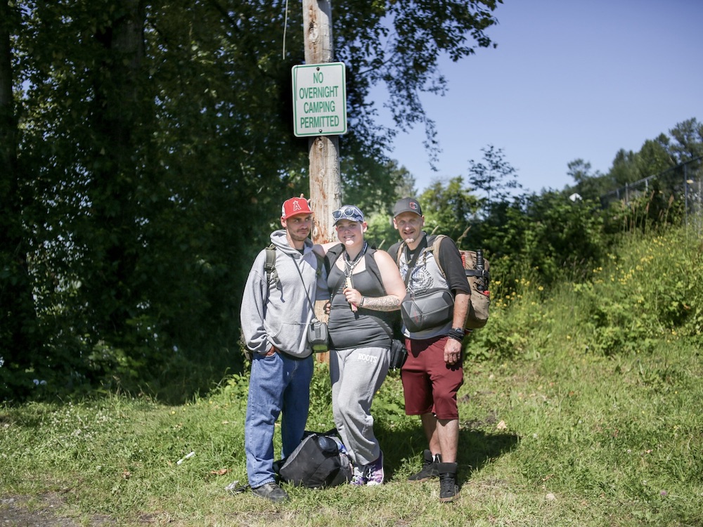 Three people stand in a grassy area in front of a telephone or hydro pole. On the pole, there is a sign that reads, &ldquo;No Overnight Camping Permitted.&rdquo;