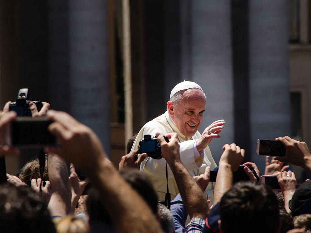 Wearing white vestments, a smiling Pope Francis holds up a hand to a crowd, many of who snap his photo with cameras and phones.