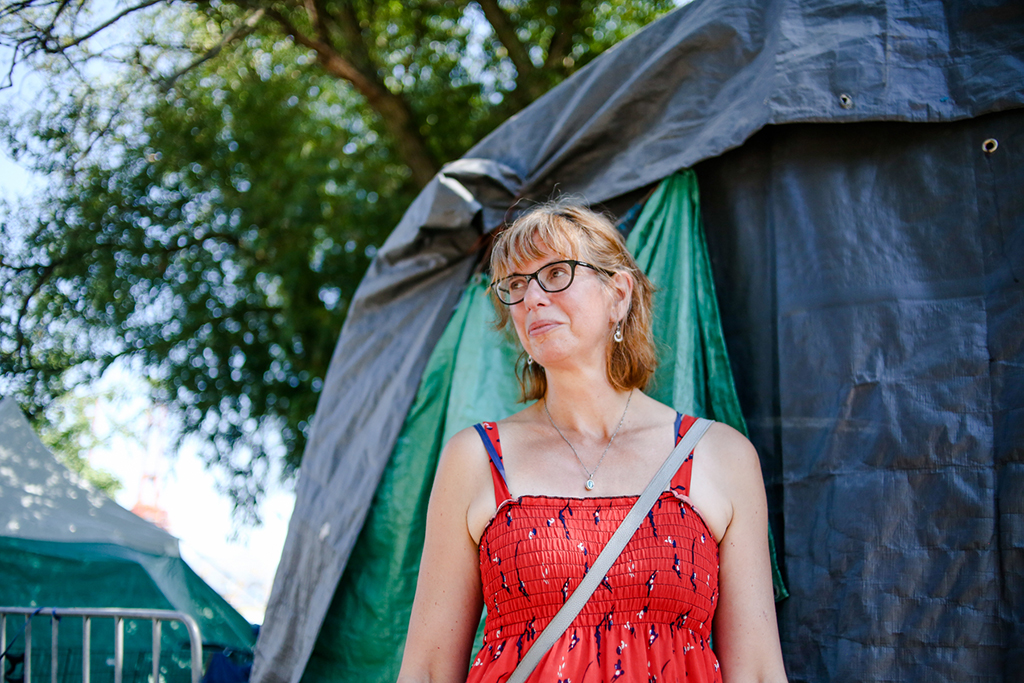 It&rsquo;s a hot day, and a woman in a red dress stands in front of a large tent in the shade of a tree. There are more tents in the background.