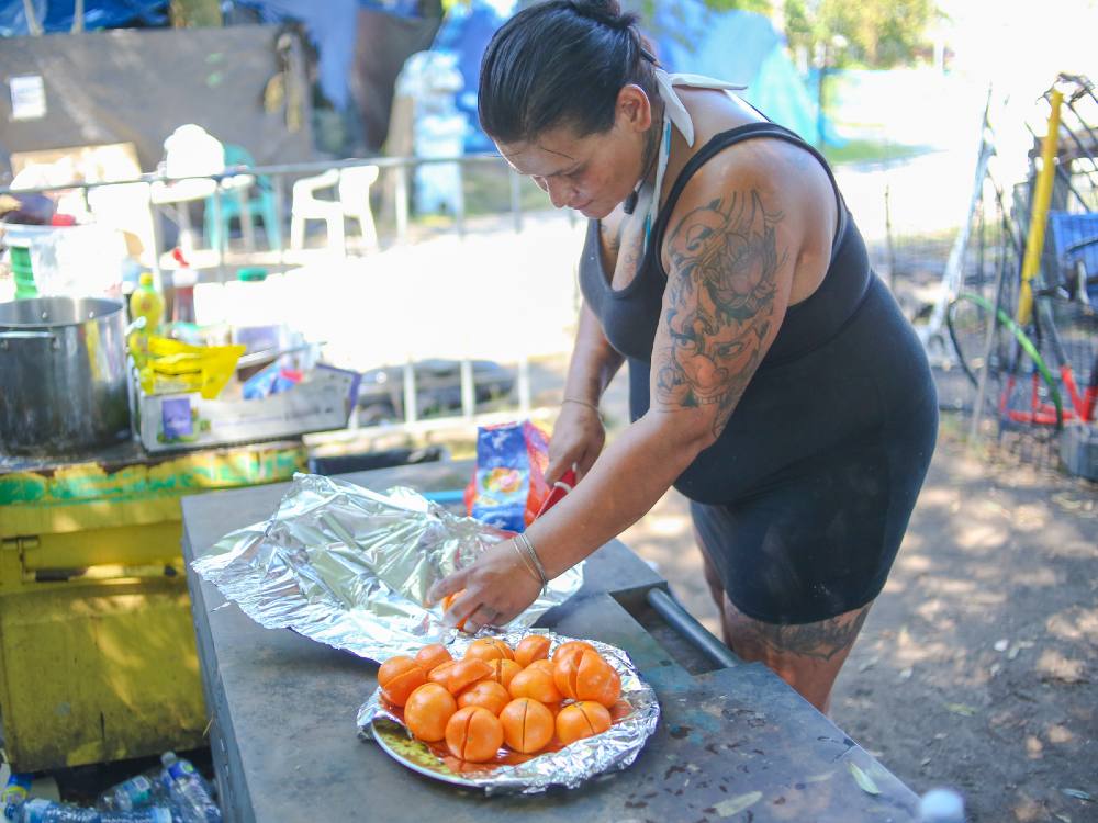A woman with a red ceramic knife halves oranges and adds them to a plate. She&rsquo;s standing in an outdoor kitchen in a park under the shade of a tree.