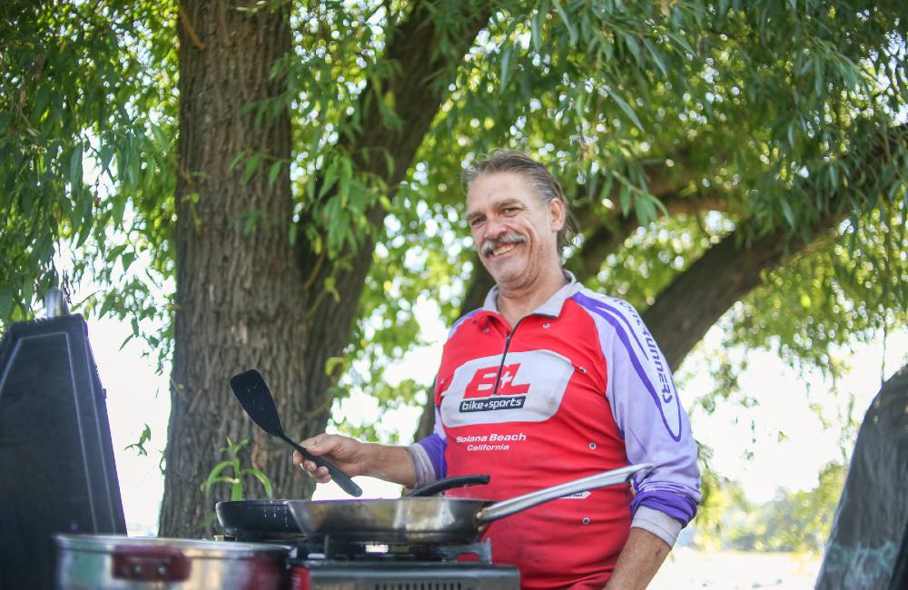 A man in a bike shirt with greying hair and mustache has a big smile on his face. He&rsquo;s holding a spatula in front of an outdoor stove under the shade of a tree.