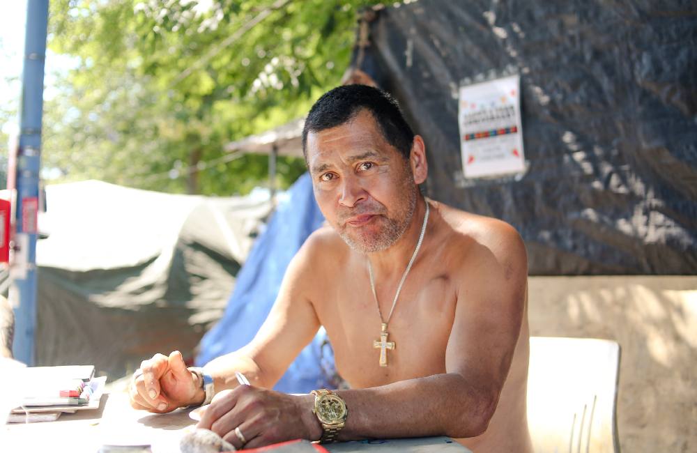 An Indigenous man with short hair, no shirt, a gold watch and a gold cross around his neck looks into the camera. He&rsquo;s sitting at a table in the shade on a hot day.