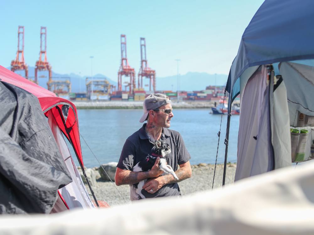A man wearing sunglasses and a cap is standing between two tents with a dog in his arms. Behind him is the water of the inlet, the red cranes of a nearby and Vancouver&rsquo;s North Shore mountains.