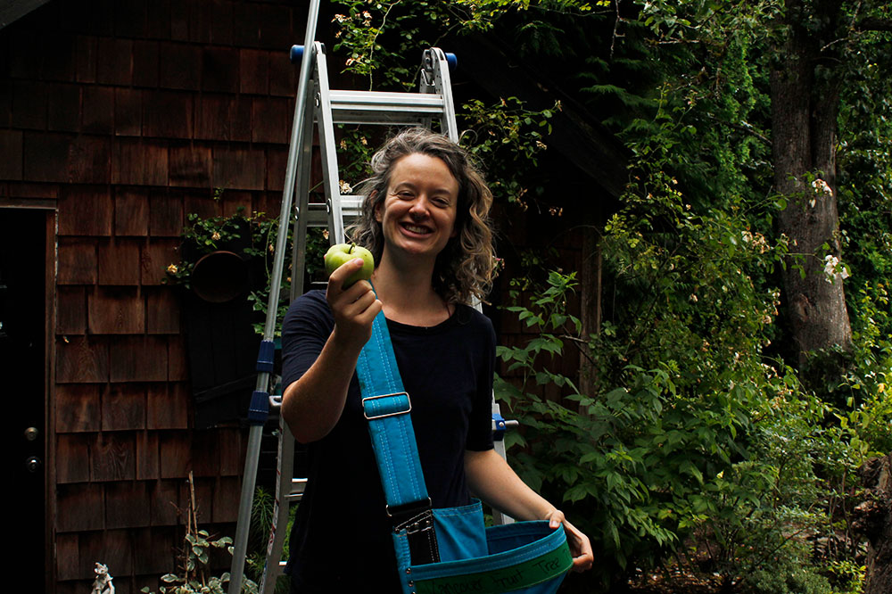 Michelle Gamage stands in front of a ladder, wearing an apple-picking bag and holding a green apple out towards the camera.