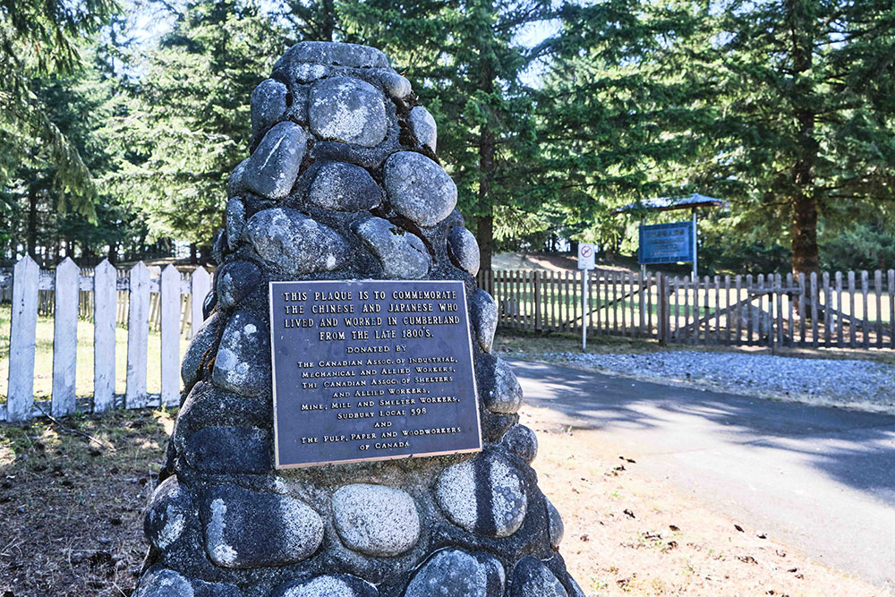 A memorial monument made of stone is mounted with a plaque that reads &ldquo;This plaque is to commemorate the Chinese and Japanese who lived and worked in Cumberland from the late 1800s.&rdquo; The entrance for the Chinese cemetery is in the background to the right.