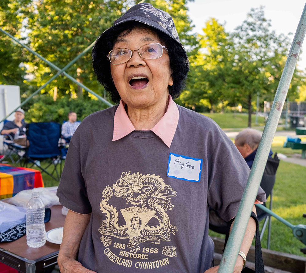 May Gee is wearing a bucket hat and smiling. She is showing off a dusty blue shirt emblazoned with a gold dragon logo that reads &ldquo;Cumberland historic Chinatown, 1888-1968.&rdquo;