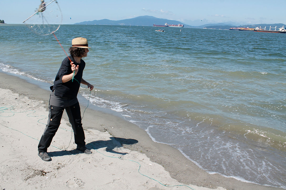 A woman in a straw hat swings a half-moon crab trap around on a red rope, preparing to throw it into the calm waters off a sandy beach.