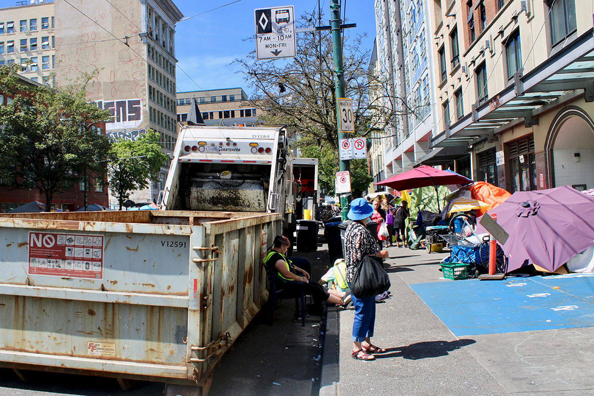 Tents, umbrellas and people&rsquo;s belongings line the sidewalk on East Hastings Street in front of buildings. City workers in reflective vests squat in front of a dumpster and a garbage truck.