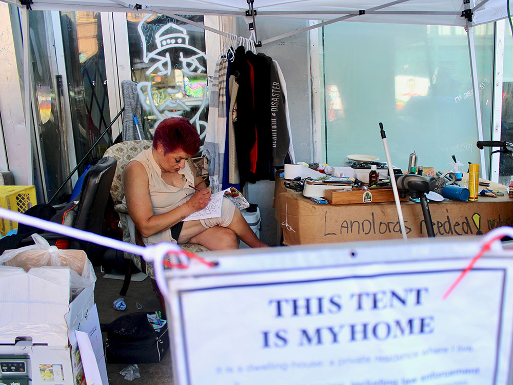 A woman with short red hair sits and colours in a doorway on East Hastings Street. Her possessions are around her and clothes hang on a makeshift rack. In front is a sign that says &ldquo;This tent is my home.&rdquo;