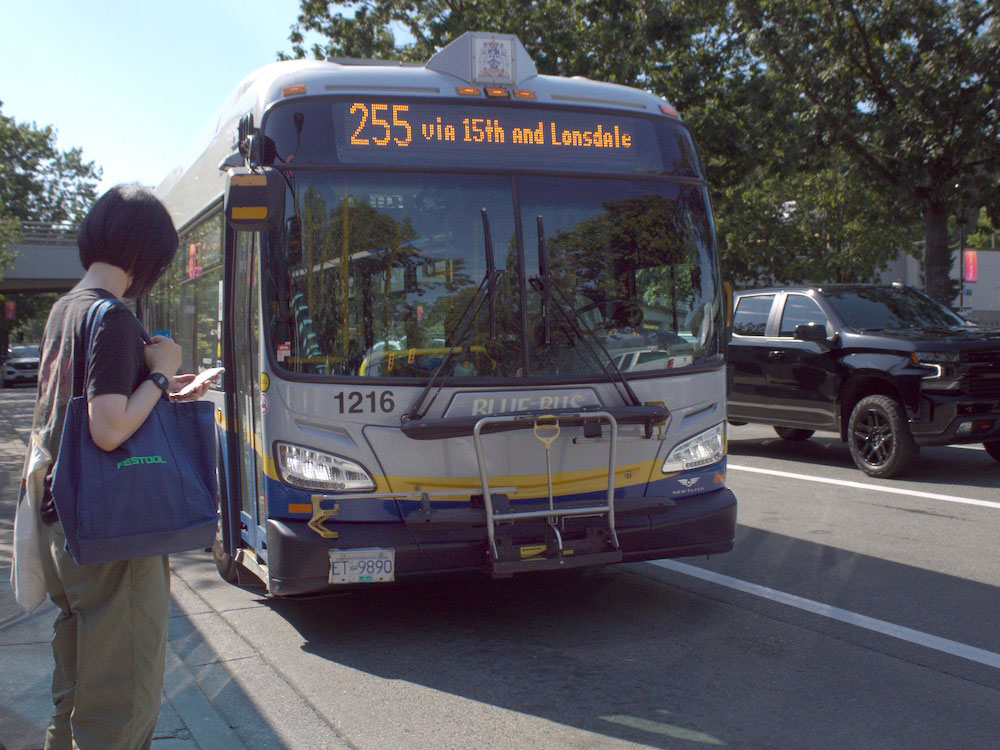 A bus with a sign that reads &ldquo;255 via 15th and Lonsdale&rdquo; slows for a passenger waiting to board to the left of the frame. The bus is blue and grey with a yellow digital sign. The waiting passenger has short black hair and is carrying a blue tote bag.
