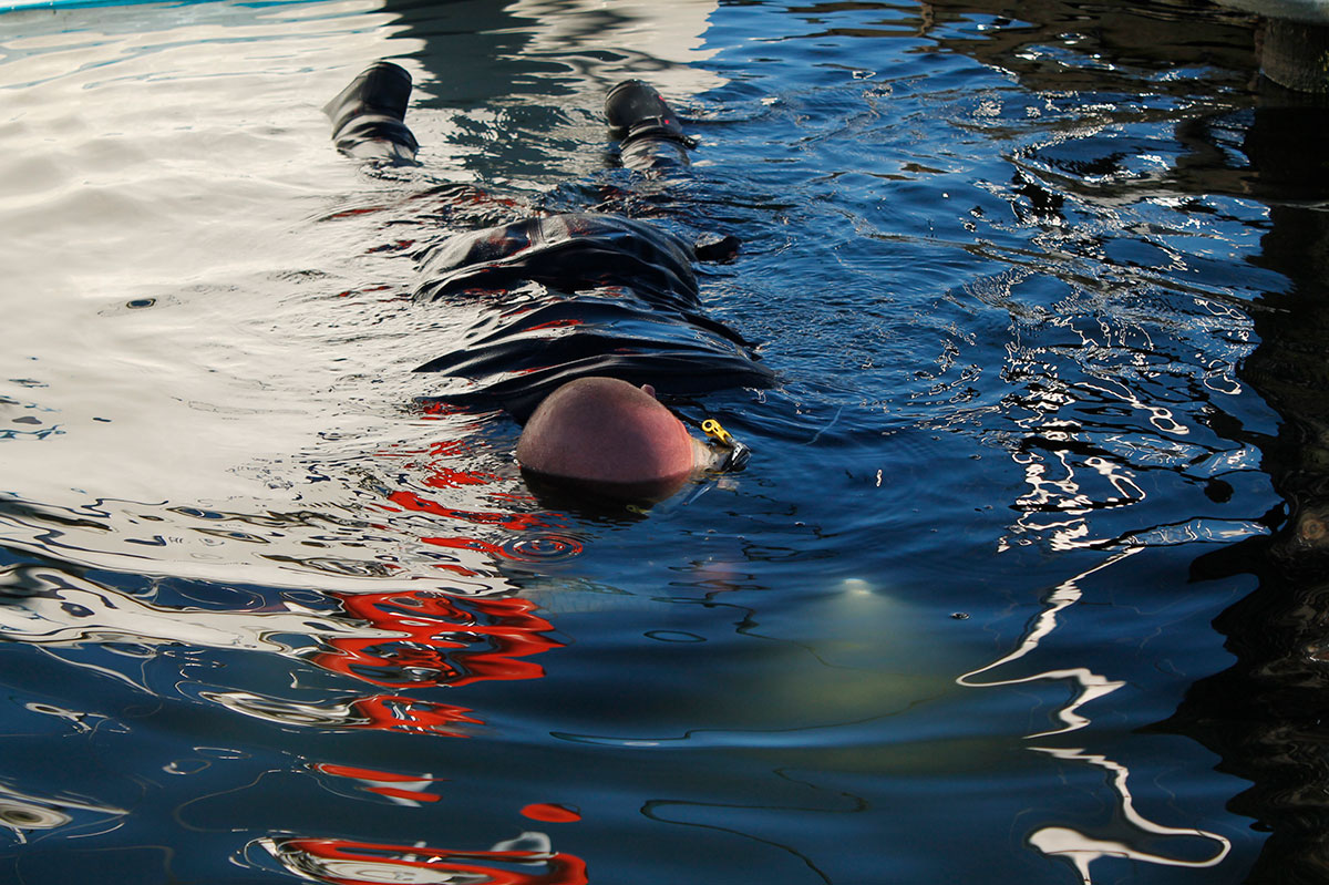 A man lies face-down in inky waters of a marina. The lights of his underwater camera shine through the water. Bright sunny reflections distort the water&rsquo;s surface. 