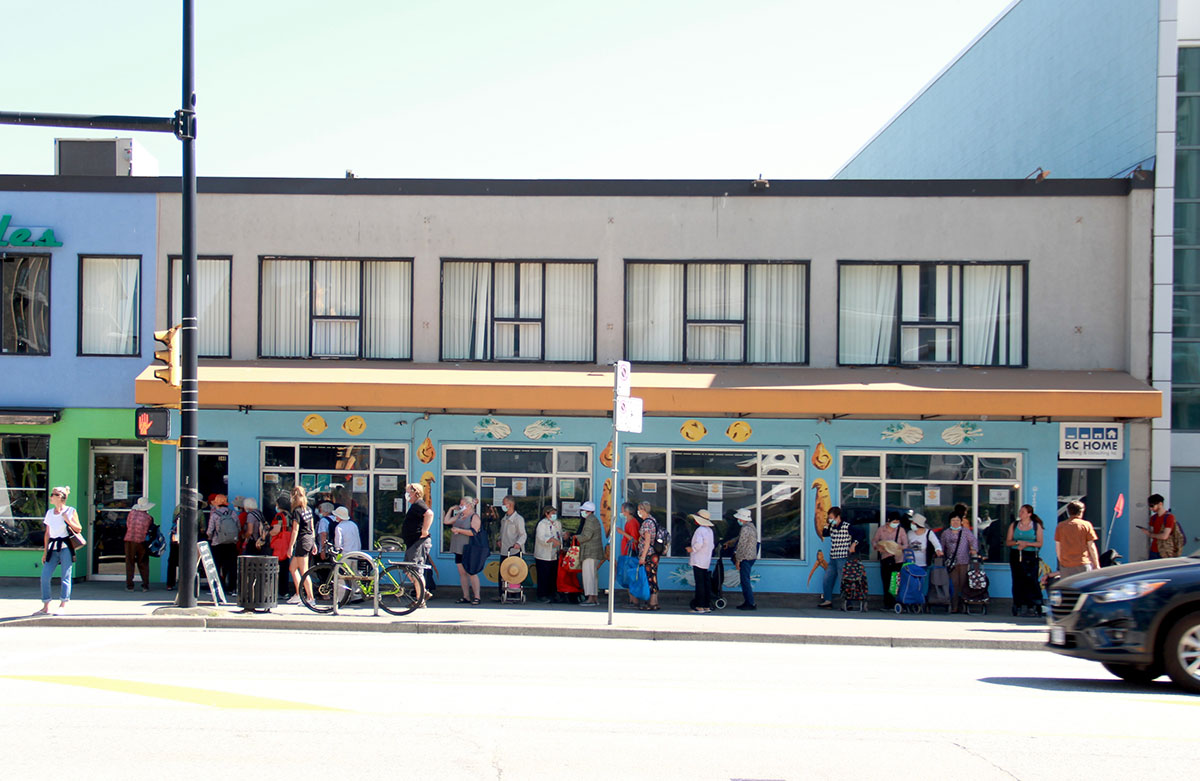 Two dozen people line up in front of the brightly painted walls waiting to access the rescued food market. They stand in the shade of a small orange awning, but the hot sun had taken over most of the sidewalk. There are no trees or grass, just cement and a road.
