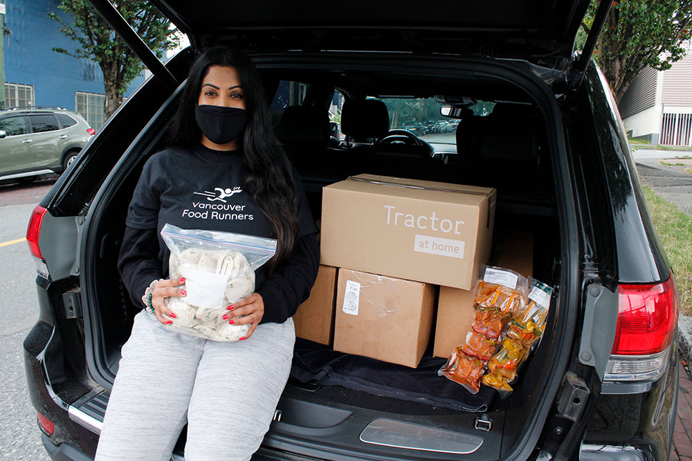 A woman wearing a face mask leans against the back of her vehicle with boxes of food from the Tractor kitchen. She’s holding a bag of cooked chicken which will go on to feed people in her city, likely that night.