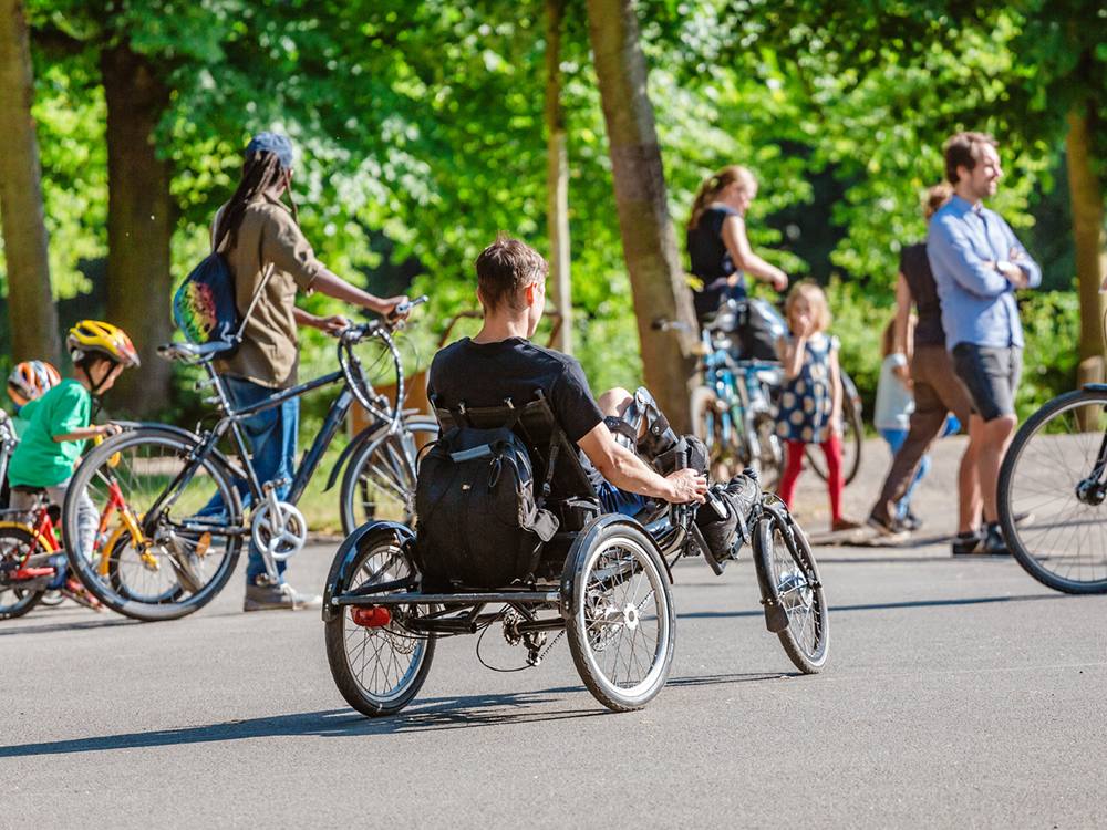 People gather on a paved path in a park on bikes. There are kids and adults. One man is using a hand-propelled bike.