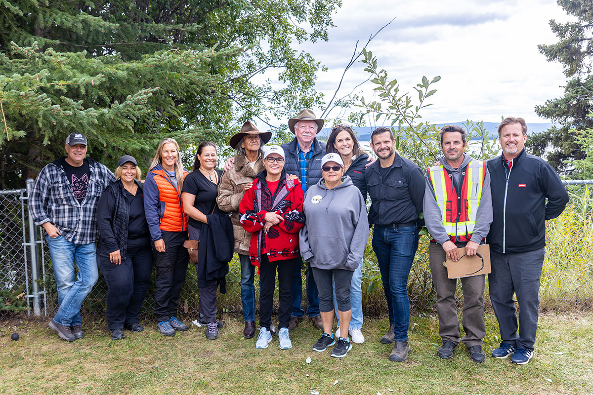 Twelve people stand in front of a fence, mostly smiling; beyond them, in the background, is a large body of water.