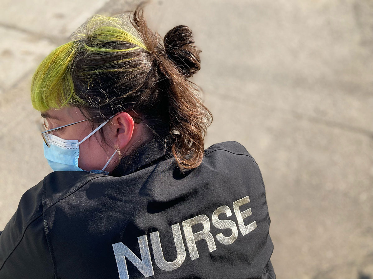 A person facing away from the camera wears a black jacket that reads &lsquo;NURSE&rsquo; in bold white letters across the back. Their face is turned to the side, showing green bangs, a medical mask and glasses. There is faded pavement in the background.