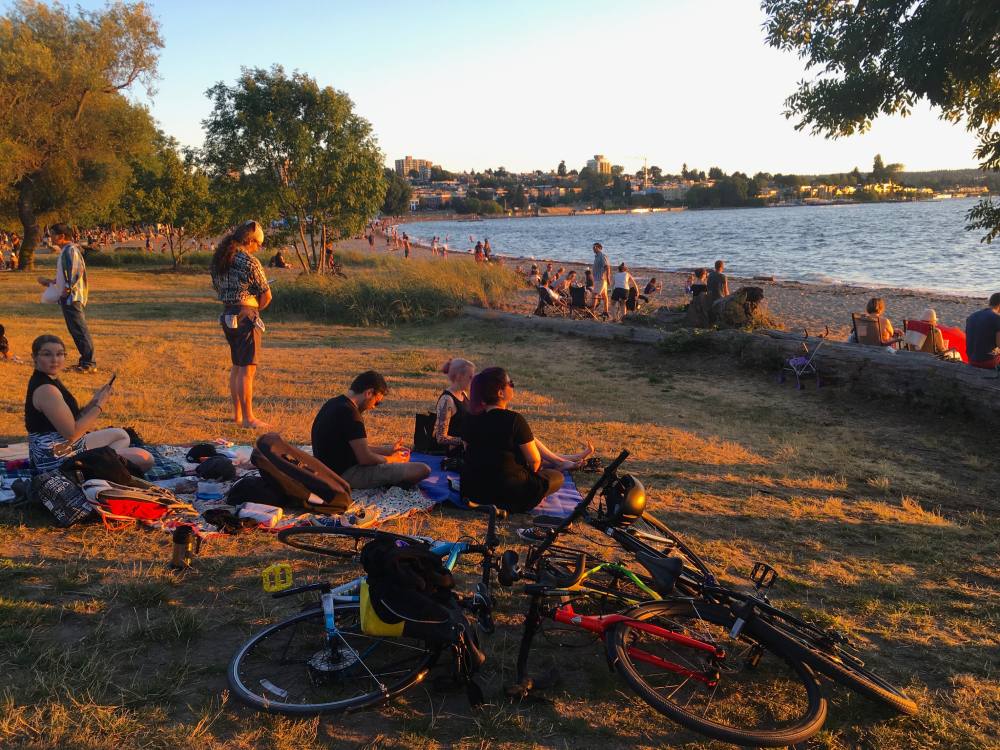 People are enjoying the sunset on picnic blankets at Vancouver&rsquo;s Kitsilano Beach. They&rsquo;ve ridden their bikes here, which are laying down in the sand.