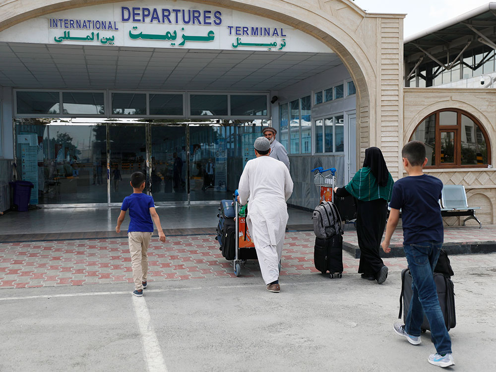 A family approaches the glass doors of a building. Above the doors, a sign reads in English and Arabic, &ldquo;International Departures Terminal.&rdquo;