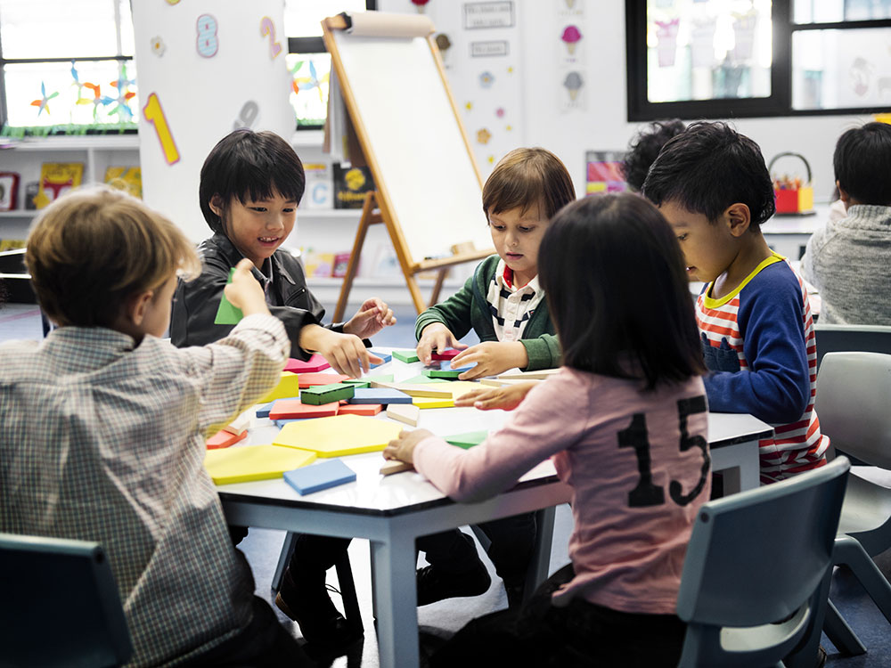 Kindergarteners sit around a table in a classroom, playing with wooden blocks.