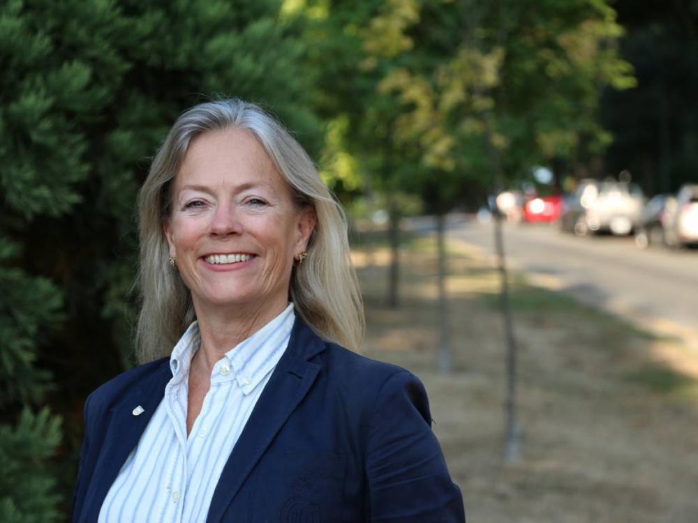 A woman with blonde hair, wearing a blue blazer, stands on a sidewalk on a street lined with trees.