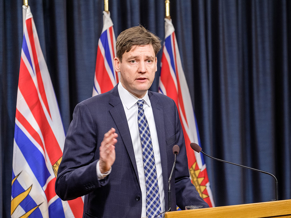A man with tousled hair, wearing a blue suit, white shirt and blue and white patterned tie stands in front of three BC flags.