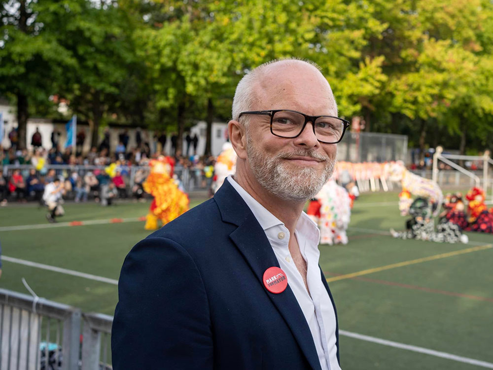 A man with short greyish hair and a neatly trimmed beard, wearing black-rimmed glasses, a white shirt and blue blazer faces the camera.