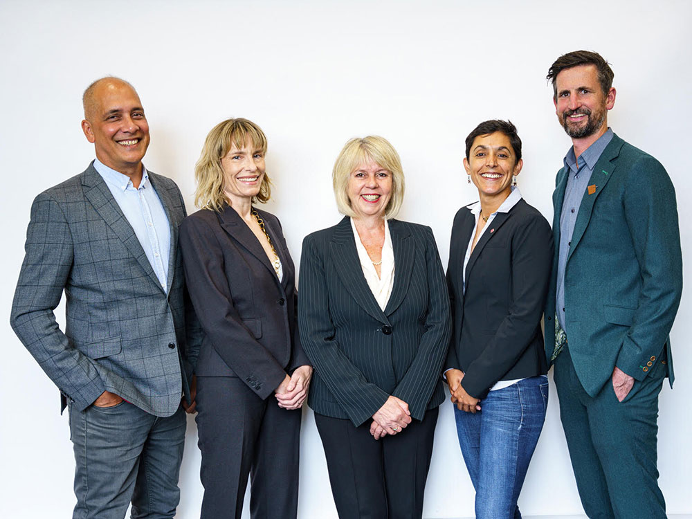 A lineup of five people dressed in business attire stand in front of a grey background. They shortest is in the centre, with people assembled outwards in height to the right and left, with the tallest people on the end. They appear enthusiastic and beaming.