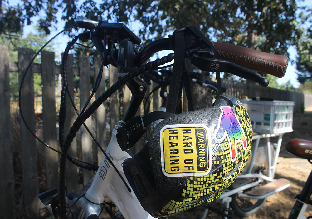 Naomi Wilde&rsquo;s bike helmet hangs on the handlebars of her white bike. The helmet is adorned with stickers, including one in yellow and black text that reads 'Warning: hard of hearing.'