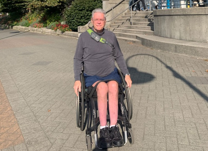 Jacques Courteau is wearing a grey shirt and shorts. He is in his wheelchair on Vancouver&rsquo;s downtown seawall. He has grey hair and glasses and is looking directly at the camera.