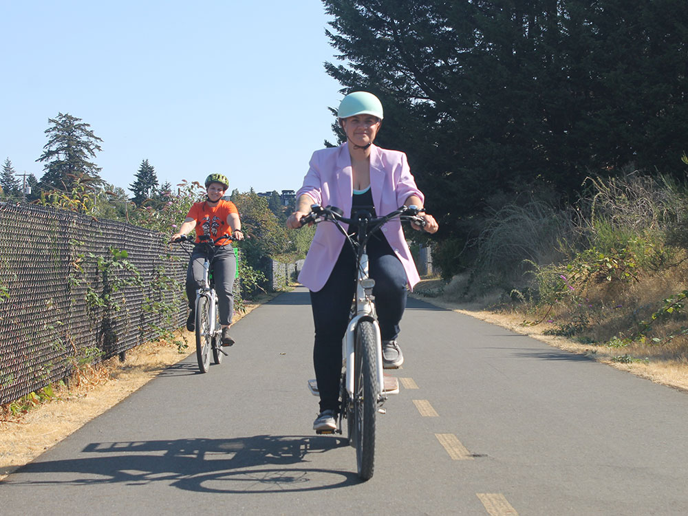 Kris Westendorp is riding her bike in the centre of the frame. She&rsquo;s wearing a lilac blazer. Her coworker Naomi Wilde is wearing an orange t-shirt, riding behind Westendorp to the left of the frame. They are on a protected bike lane and smiling.