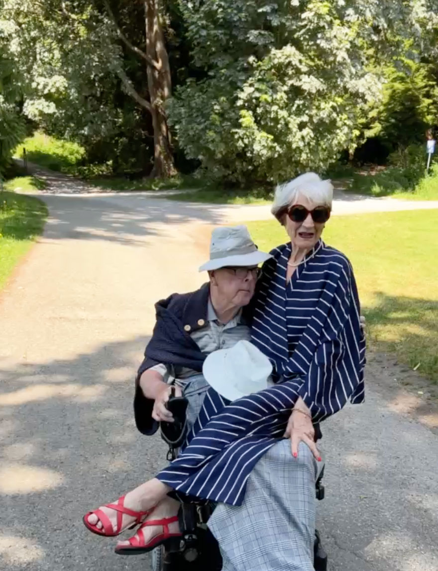 Peter and Janice Foran ride down a protected bike lane. Janice is wearing a navy dress and sunglasses, sitting on Peter&rsquo;s lap. Behind them are trees and green grass.