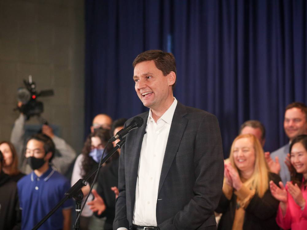 A smiling man with short dark hair, wearing a suit with a white shirt and no tie, stands behind a podium and microphones with supporters and journalists in a line behind him.
