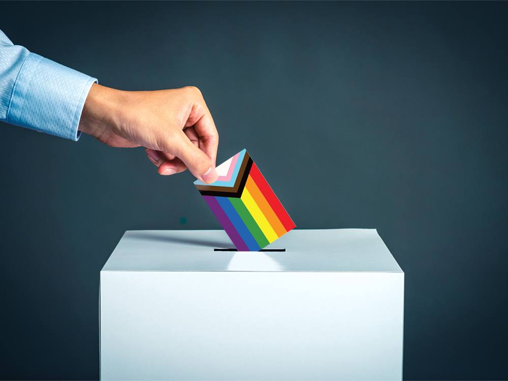 A person’s hand drops a ballot into a voting box. The ballot depicts a miniature pride flag.