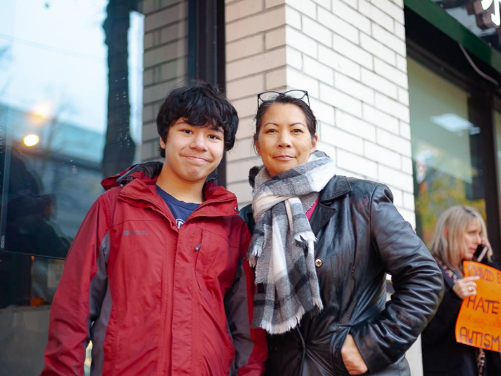 Rozann Pedersen stands to the right of the frame. She is an Asian woman in a black jacket and scarf with a pair of glasses resting on her head. Her black hair is tied back and her expression is serious but warm. Her son, Lucas, stands to the left of the frame. He has short dark hair and is wearing a bright red jacket.