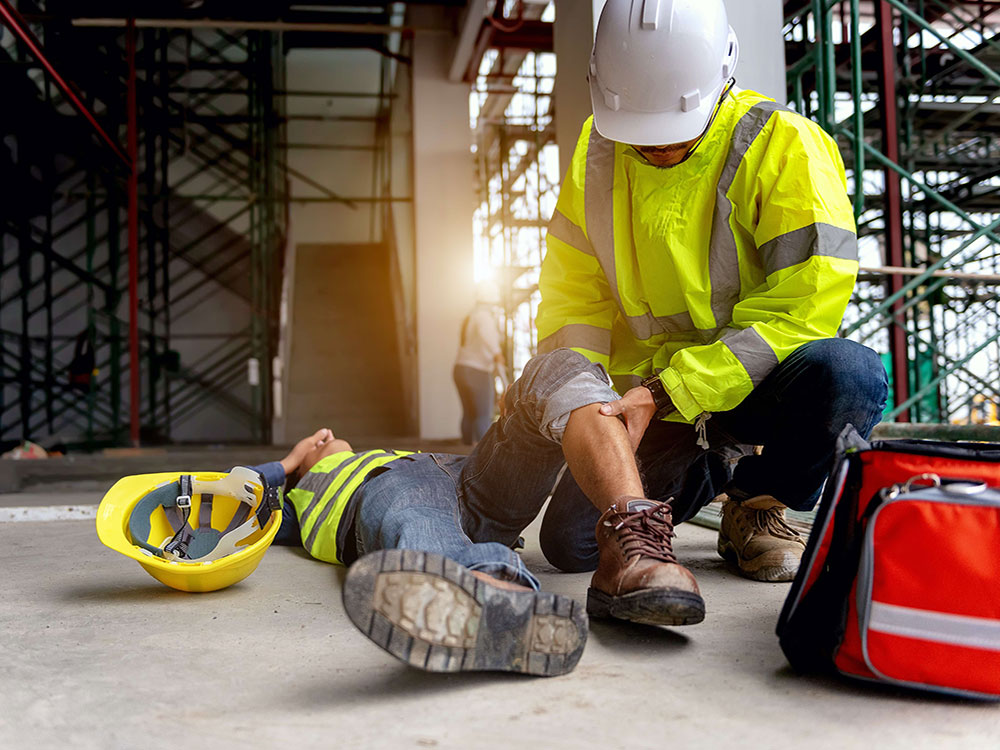 A person lies on the ground on a construction site with their knee in the air, while another person attends to them. Both people are wearing PPE. 
