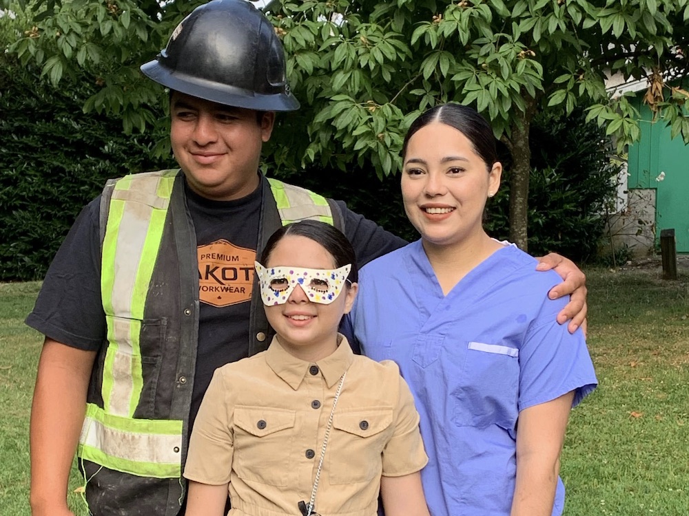 A family of three poses together for a photo. On the right stands Claudia Zamorano in a blue hospital uniform.