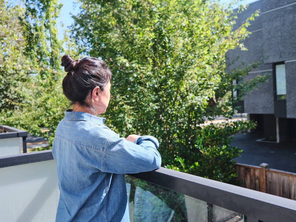 A Chinese woman stands on a balcony overlooking a crop of trees and a neighbouring grey building. It&rsquo;s a sunny day. The woman is turned away from the camera and she is resting her arms on the balcony ledge. She is wearing a denim shirt and her black hair is tied in a topknot bun.