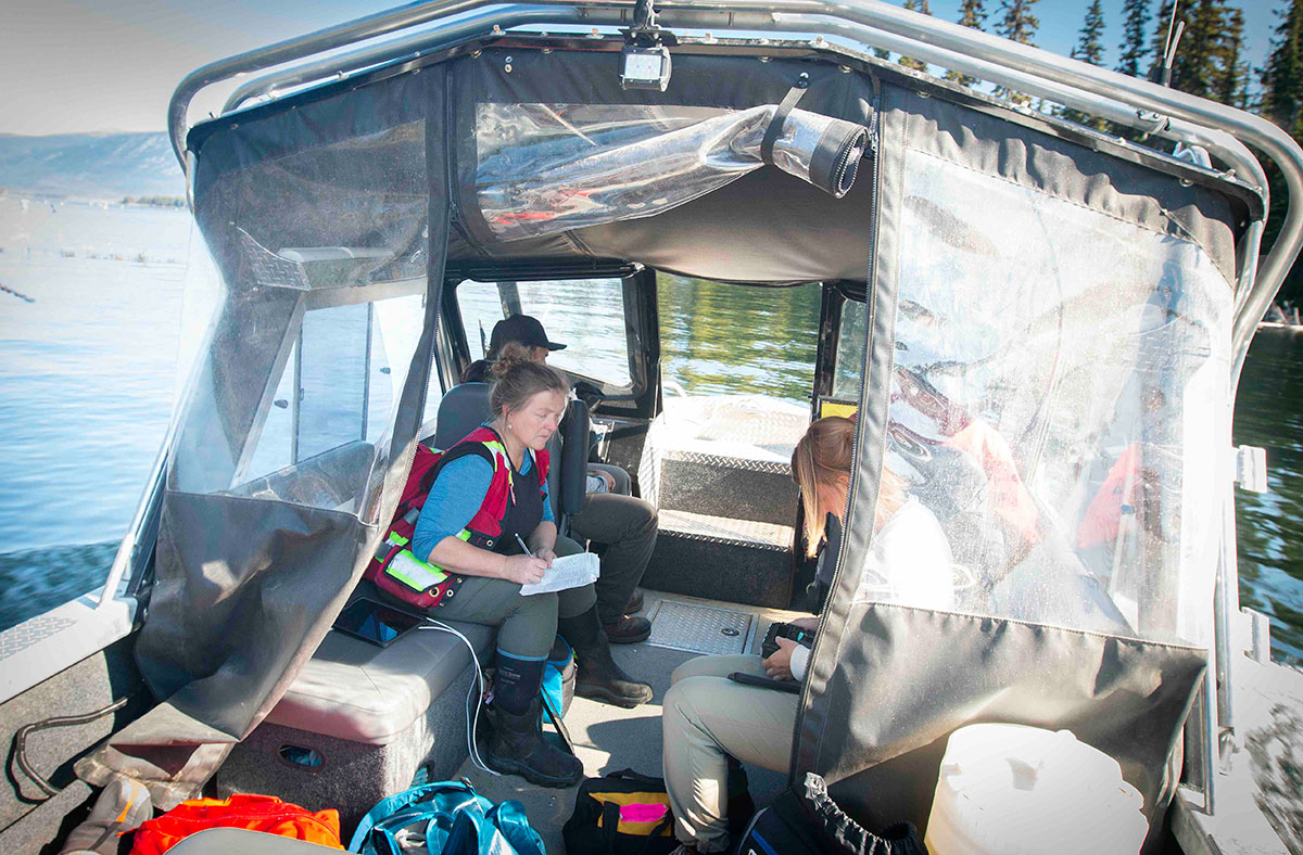 Several people sit in a boat that floats on the water. At the centre of the photo, a woman in a high-visibility vest takes notes. 