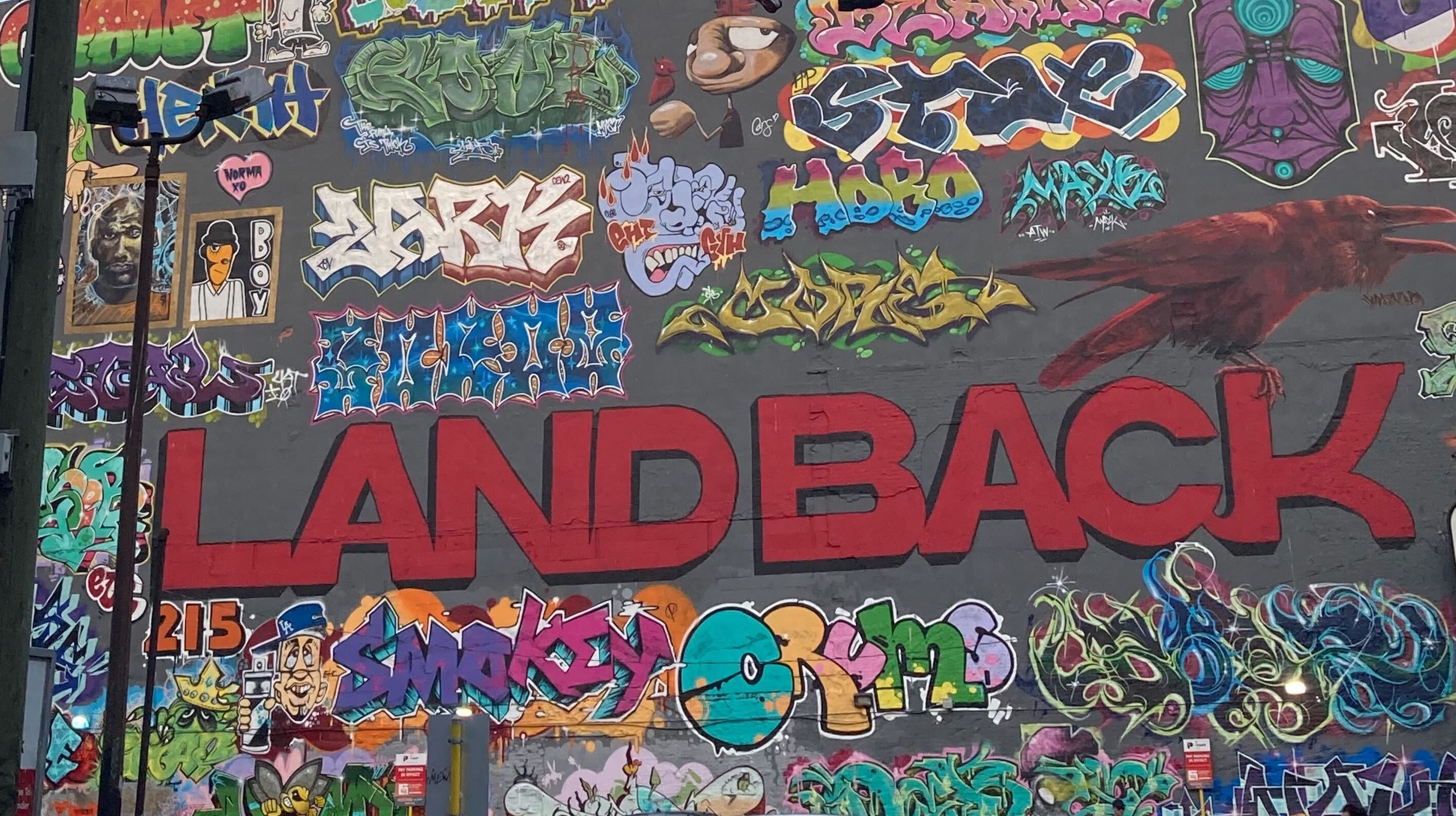 A large-format graffiti mural features numerous colourful tags from local artists unified by the words &ldquo;LAND BACK&rdquo; in large sans-serif lettering in red. The main lettering and surrounding colourful tags are on a grey charcoal painted wall.