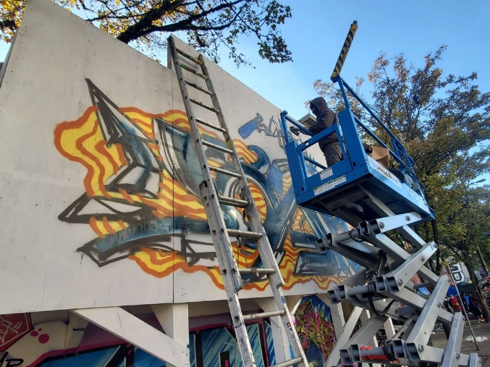 A man in a dark grey hoodie over a tuque and mask stands in a blue cherry picker while he sprays paint on a graffiti wall in Vancouver&rsquo;s Downtown Eastside. The artwork uses orange, yellow, blue and black paint. A silver ladder is in the foreground against the wall. In the background is a stand of green trees and a busy sidewalk full of people.
