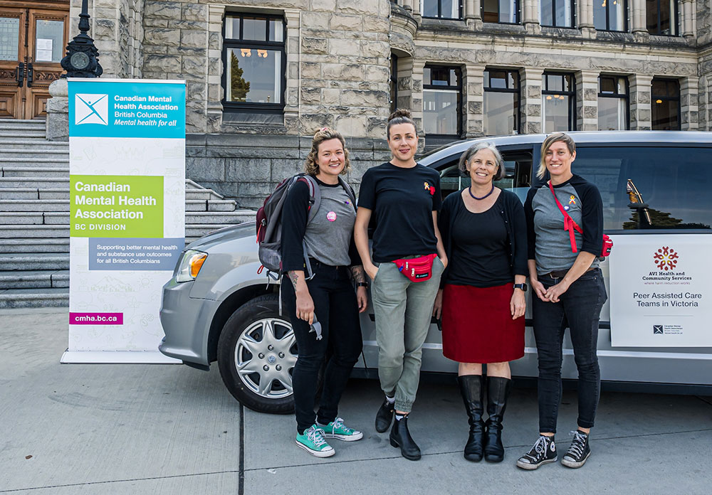 Four people stand in front of an SUV parked in front of the provincial legislature in Victoria. A large sign reads, &lsquo;Canadian Mental Health Association, BC Division.&rsquo;