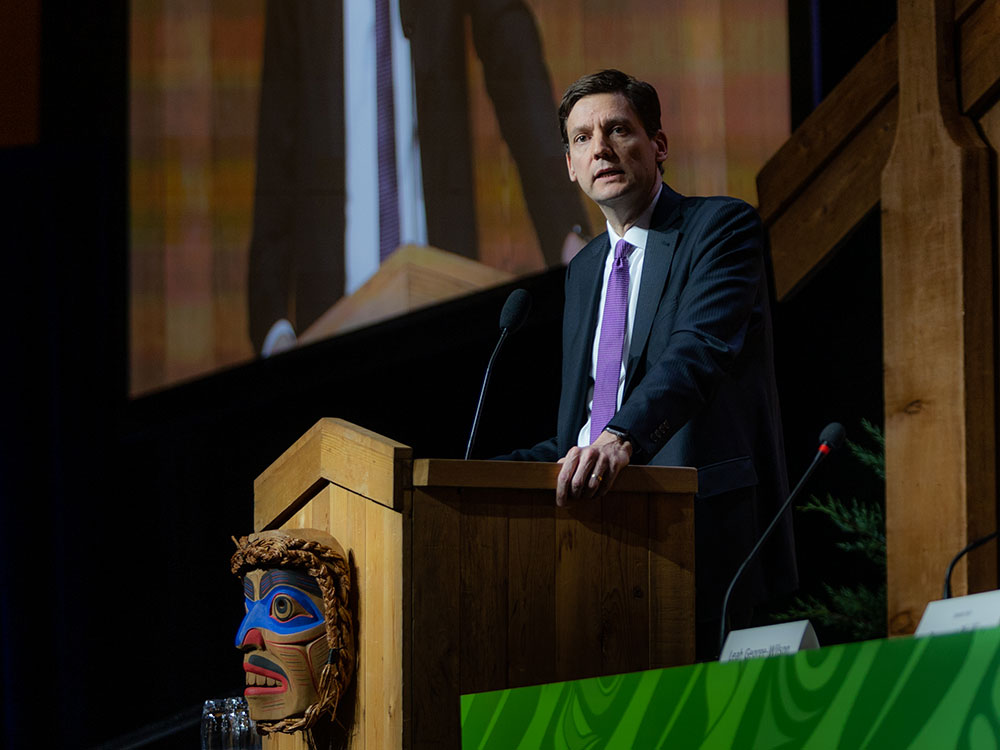A man wearing a suit and purple tie stands at a podium that has a carved Indigenous mask on the front.