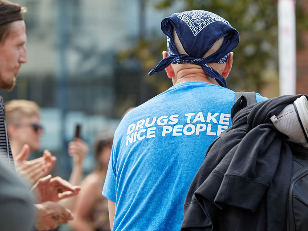 A man, viewed from behind, wears a blue bandanna, a backpack, and a T-shirt that reads, &ldquo;Drugs Take Nice People.&rdquo;