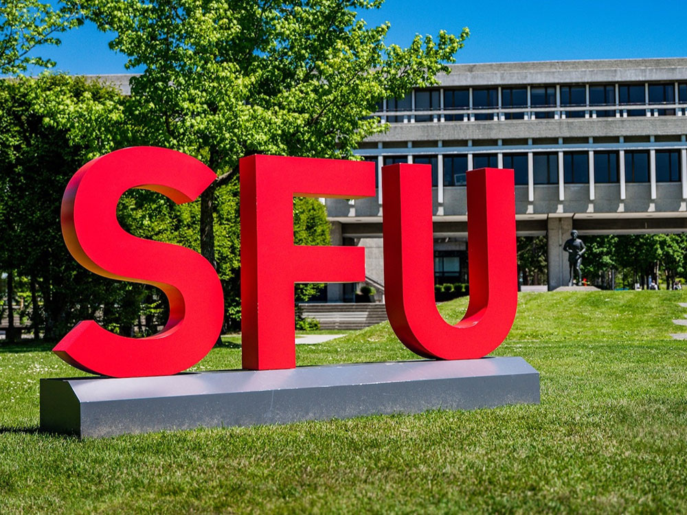 A sign consisting of bright red letters spelling &lsquo;SFU&rsquo; stands in front of a two-storey elevated concrete building.