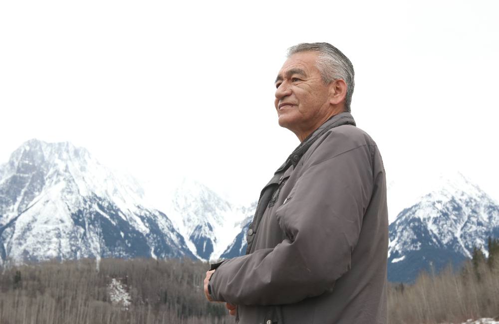 A man with greying hair, who is wearing a grey jacket, stares into the distance. There are mountains in the background.