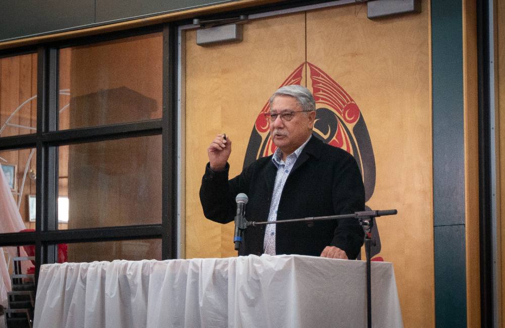 A man with greying hair wearing a black jacket and button-down shirt stands at a podium and gestures with his hand as he speaks.