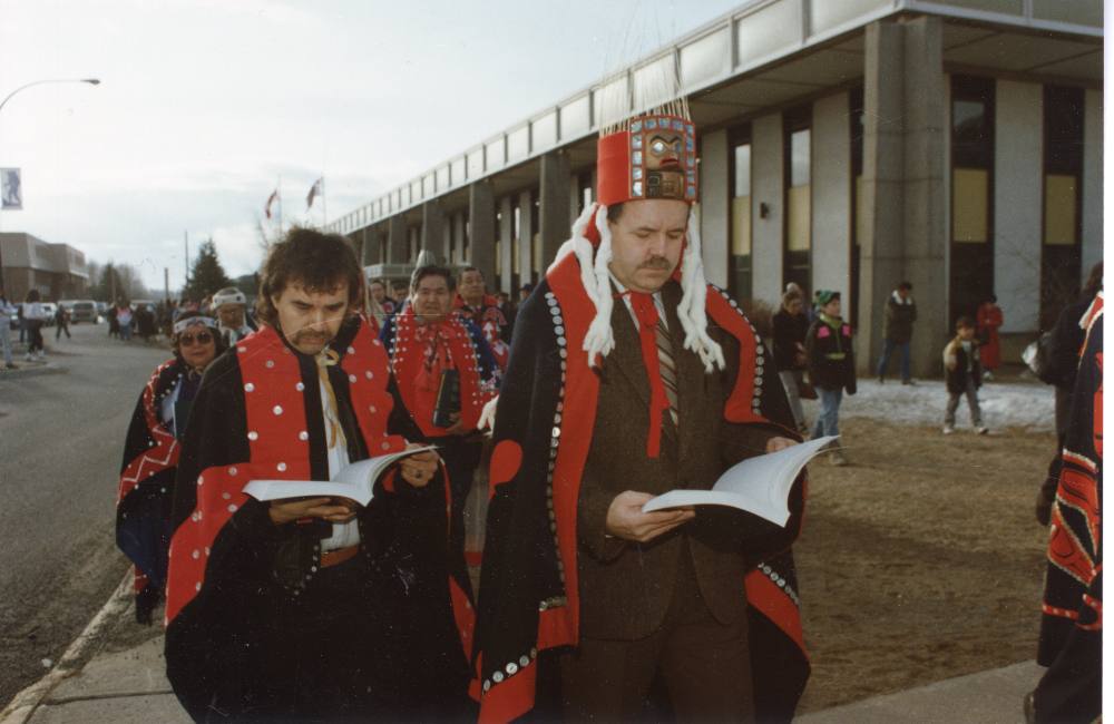 Two men look down at books they hold in their hands as they walk away from a large concrete building. Both the men and the people walking behind them wear traditional Indigenous regalia.