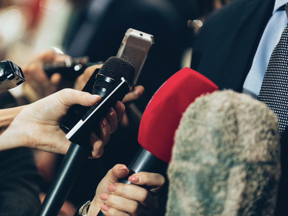 A crowd of hands holding microphones and voice recorders tilts their devices towards the right side of the screen, where a person in a navy jacket, blue shirt and dark dotted tie is standing. The hands belong to journalists in a press scrum. We can&rsquo;t see their faces. 