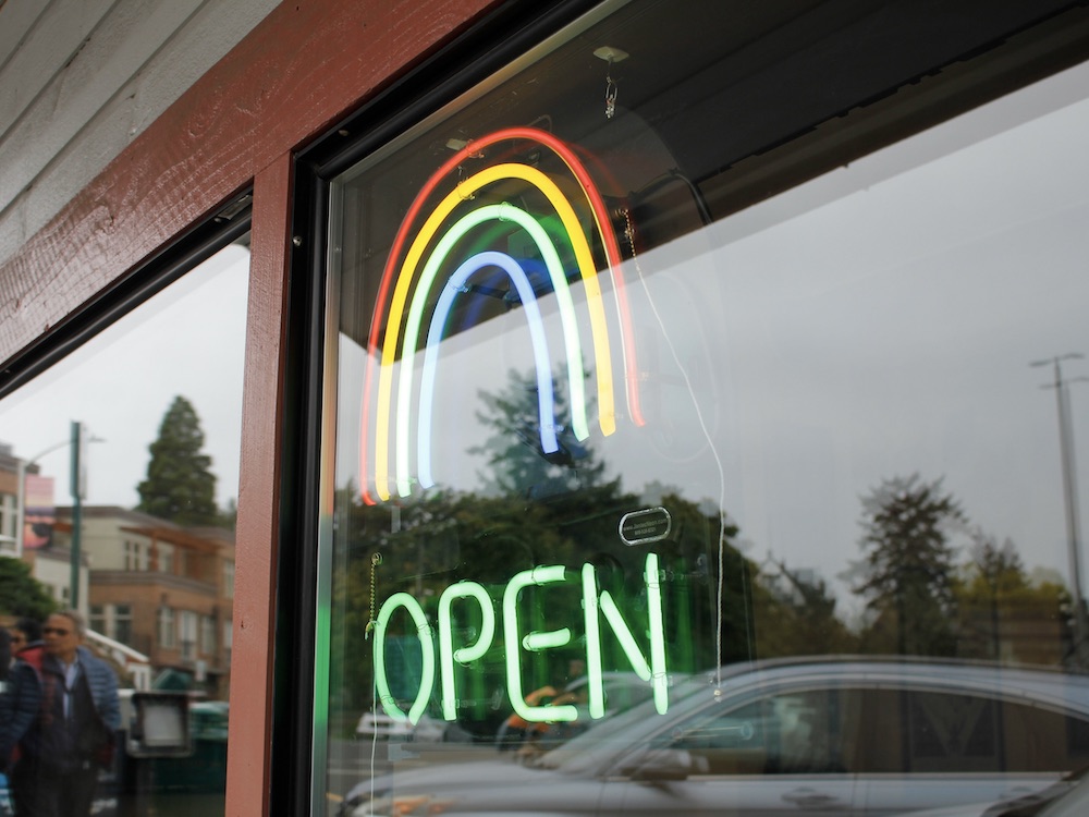 A storefront window, with a neon &lsquo;OPEN&rsquo; sign under a Pride rainbow symbol. Pedestrians and cars are reflected in the window.