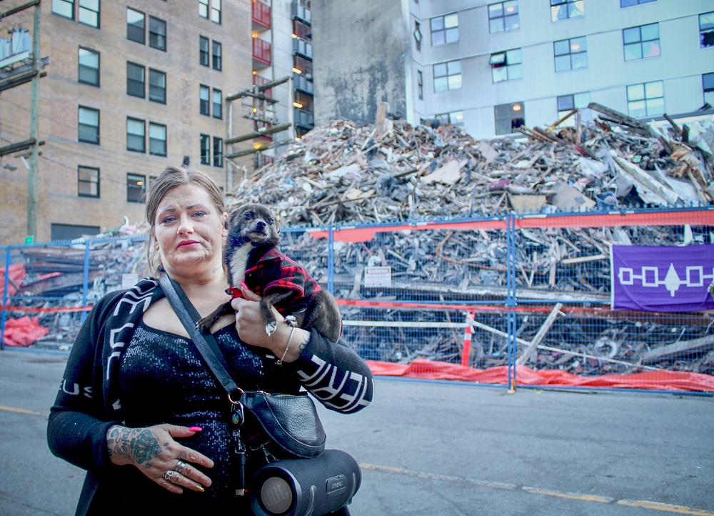 A woman holding a small dog stands on a sidewalk in Vancouver&rsquo;s Gastown neighbourhood. In the background, a huge pile of rubble from a building that was torn down after a fire sits behind a fence.